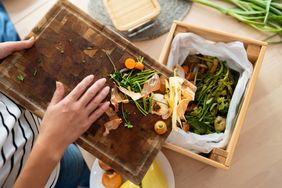 Person discarding vegetable scraps from a cutting board into a compost bin overhead view of kitchen setup