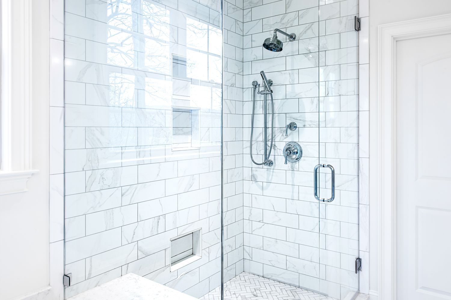 A modern glassenclosed shower with white tiled walls and silver fixtures
