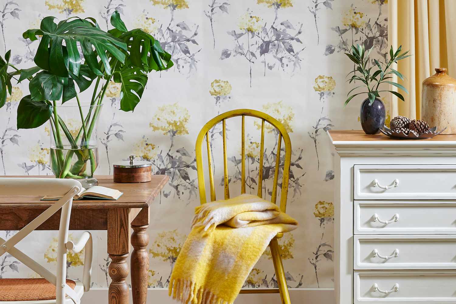 A cozy dining setup with a wooden table a yellow chair draped with a blanket and floral wallpaper in the background a dresser beside