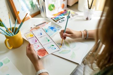 A person painting with watercolor on paper using a palette and brushes on a desk