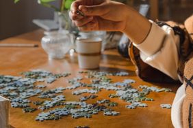 A person assembling a jigsaw puzzle on a wooden table surrounded by scattered puzzle pieces