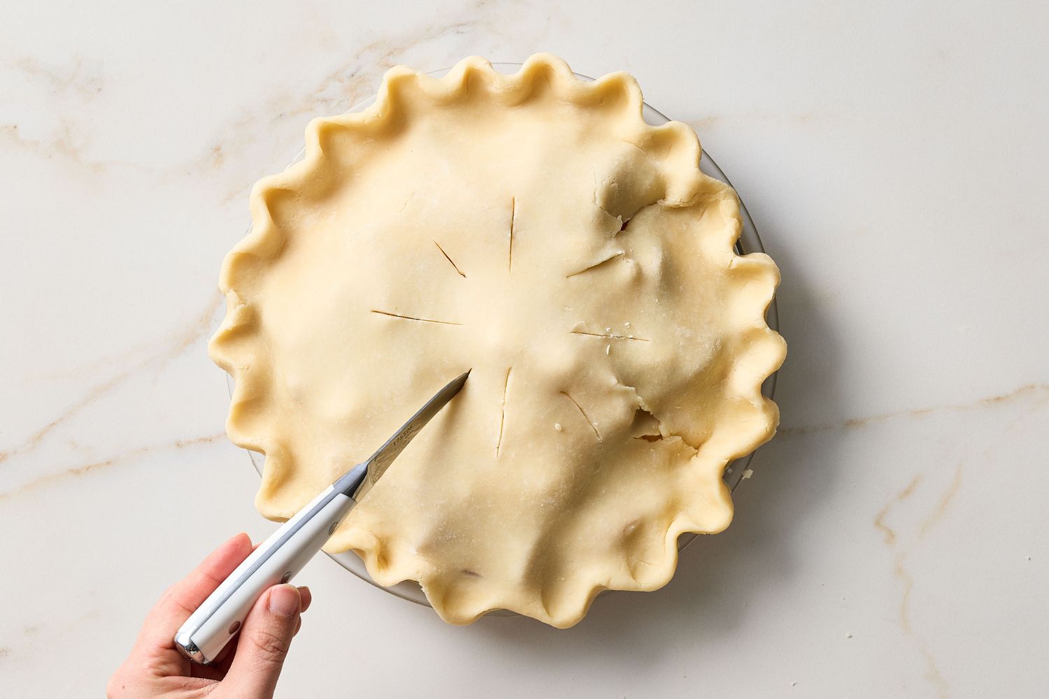 A pie with a top crust being prepared a knife creating slits in the dough