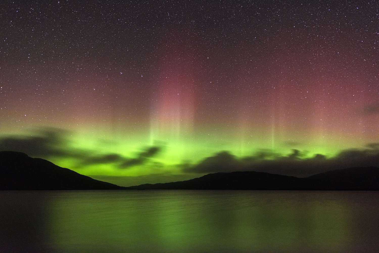Northern lights over a calm lake silhouetted hills in the background