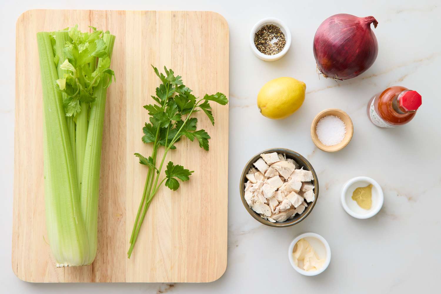 Ingredients for chicken salad on a cutting board and table, including celery, parsley, lemon, diced chicken, and condiments
