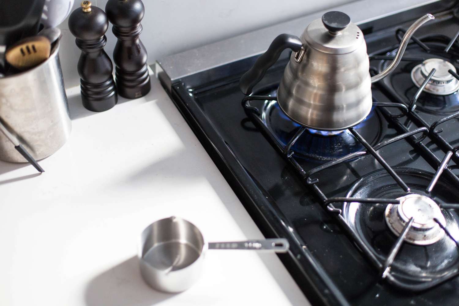 Stovetop with a kettle heating on a burner a measuring cup on a countertop nearby and kitchen utensils in a container