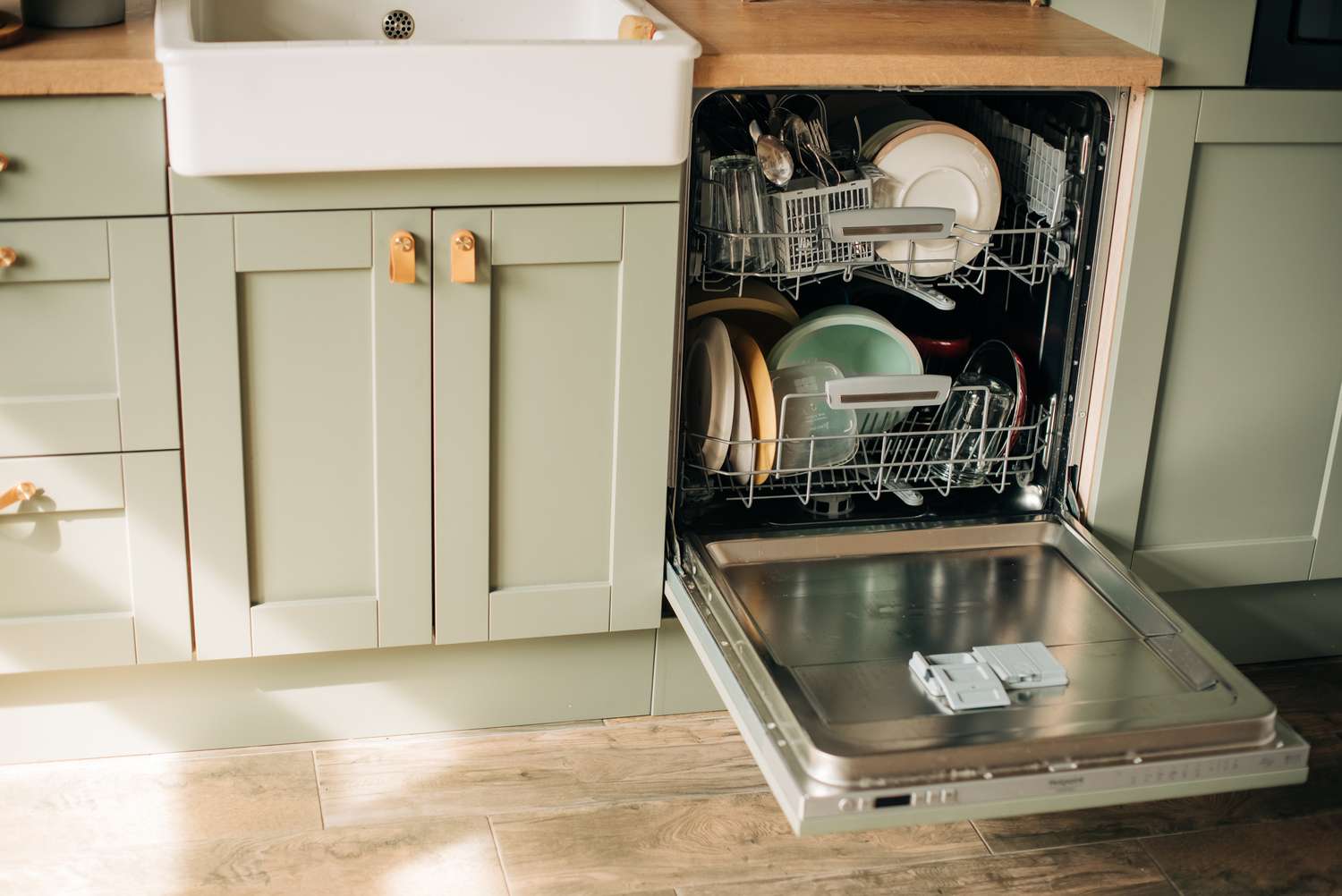 An open dishwasher in a kitchen loaded with dishes and utensils next to a sink and cabinets
