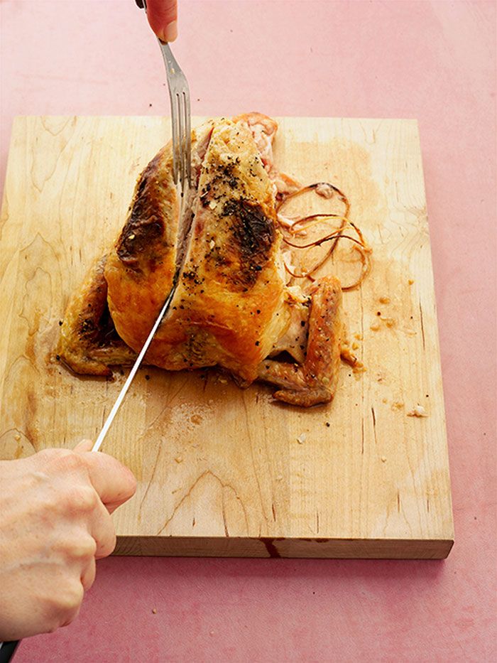 A person carving a roasted chicken on a wooden cutting board using a knife and fork