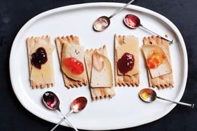 Crackers topped with slices of cheese and various fruit preserves arranged on a white plate with accompanying spoons of jam