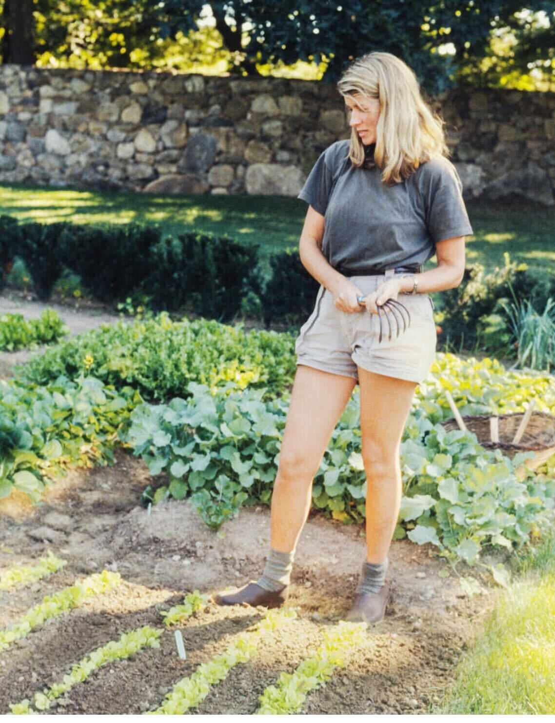 A woman standing in a garden holding a small hand rake surrounded by plants and greenery