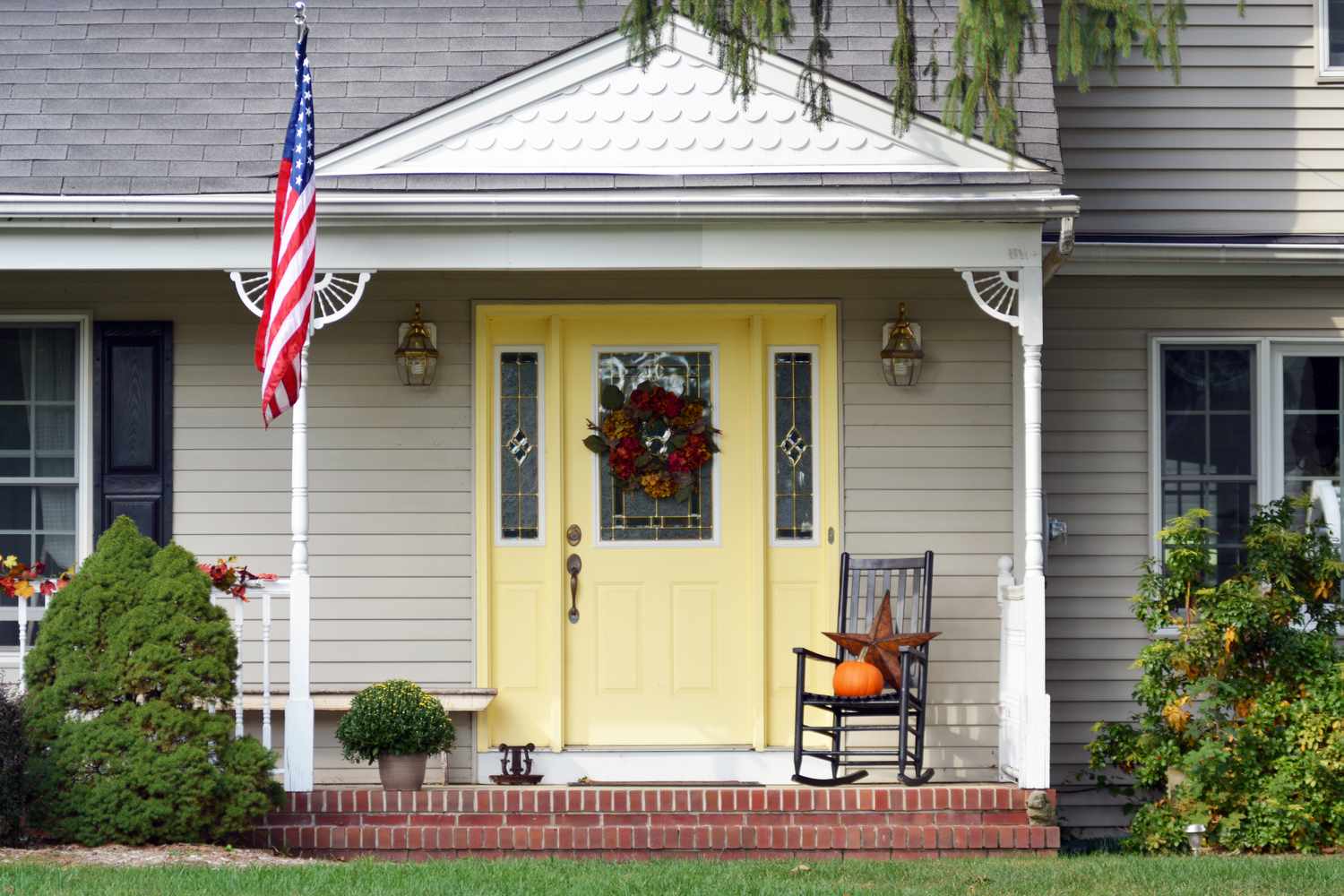Front porch of a house with a yellow door a wreath a rocking chair with a pumpkin and an American flag on the side
