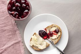 Two slices of bread on a plate one spread with butter and the other with cherry jam a knife resting on the plate jar of jam nearby