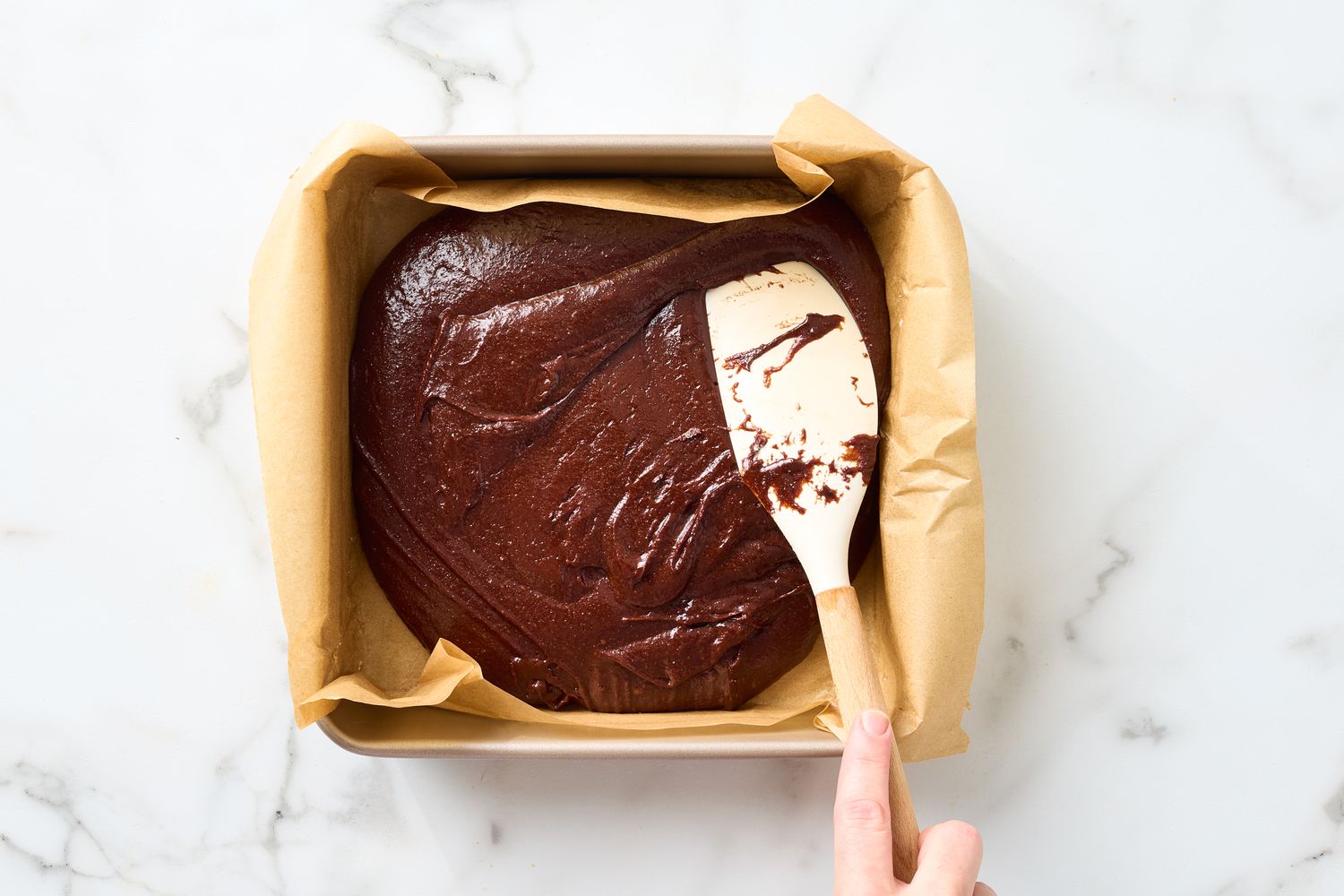 Brownie batter spread in a parchmentlined baking pan with a spatula