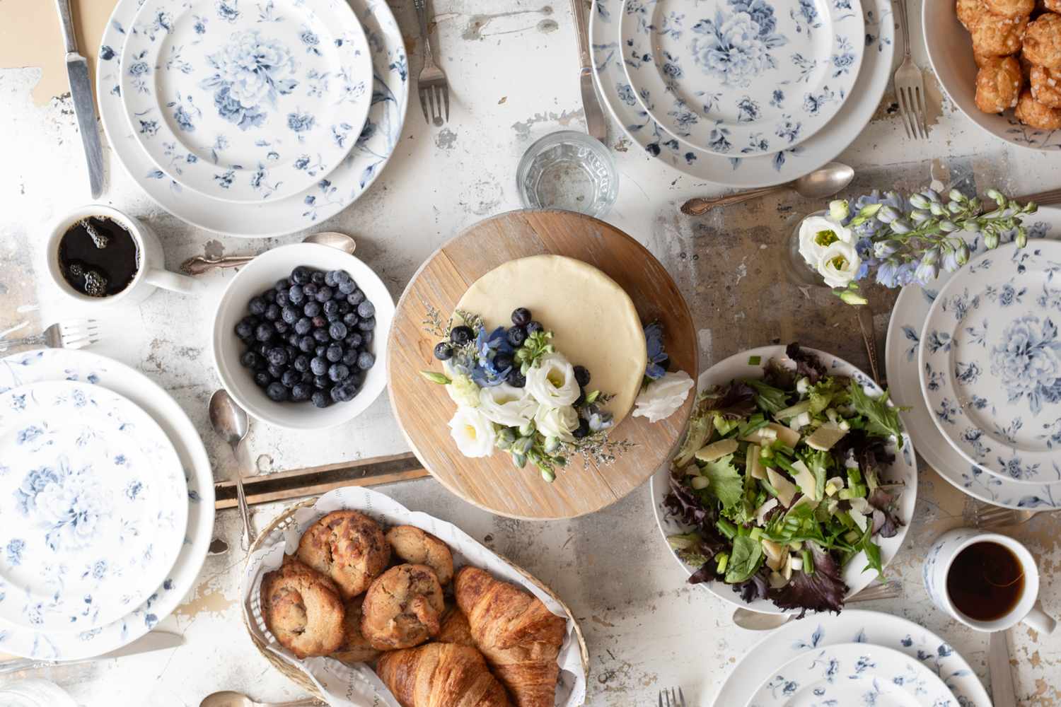 A table setting featuring floralpatterned dinnerware a centerpiece cake topped with flowers salad blueberries croissants and other dishes