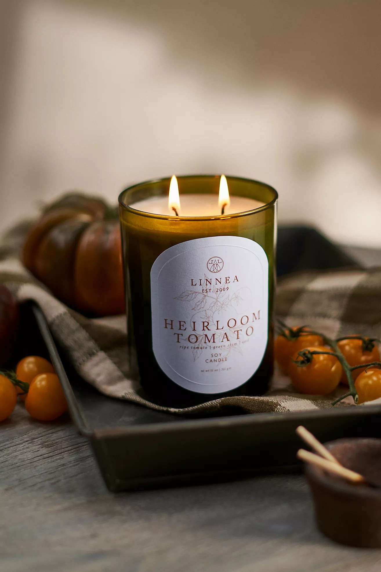 A lit candle with the label 'Heirloom Tomato,' surrounded by fresh tomatoes