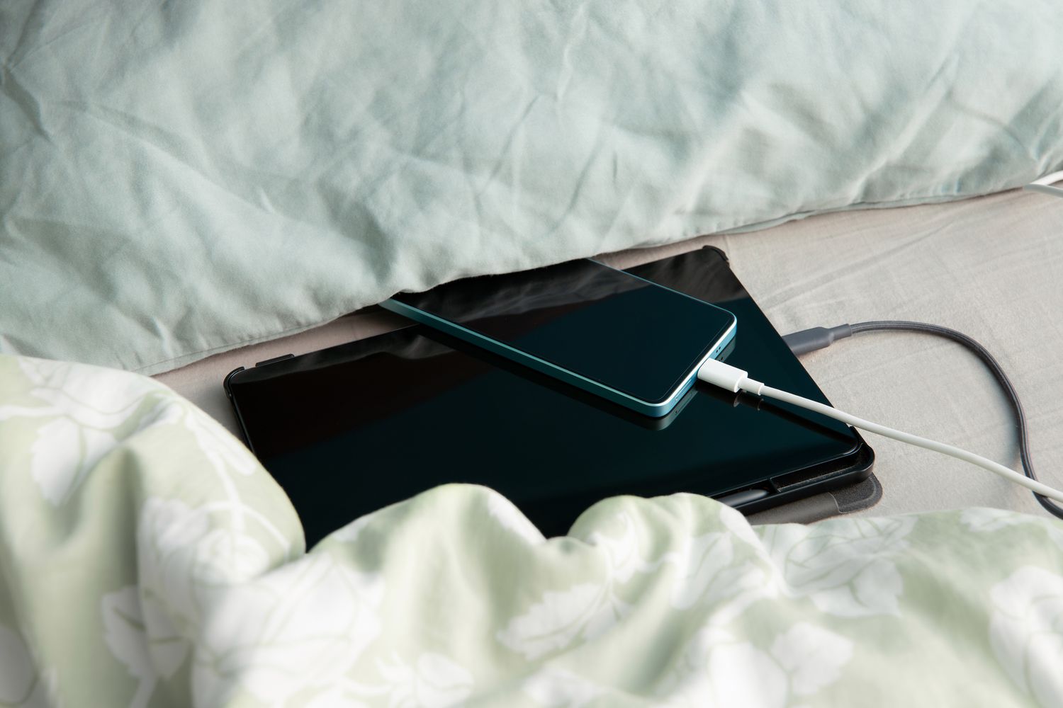 Smartphone and tablet computer are charging in bed under a pillow and blanket