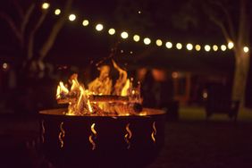 A fire pit with glowing embers and flames outdoors with string lights in the background