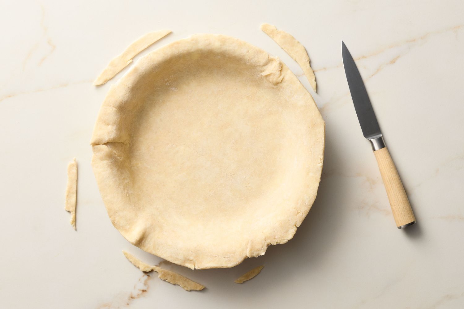 A pie crust in a pie dish with trimmed edges and a knife on a countertop