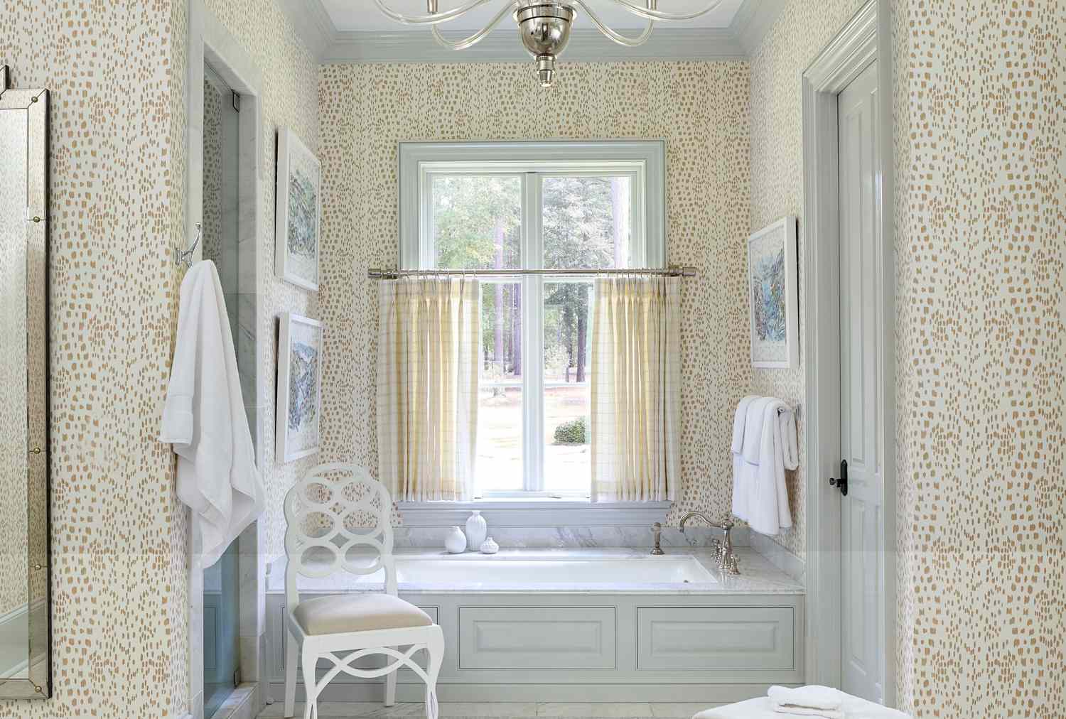 A bathroom with a patterned wallpaper a bathtub centered below a window a chair and neatly arranged towels and artwork