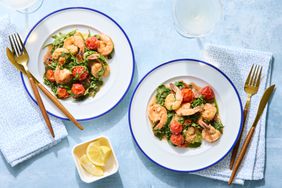 Two plates of sautéed shrimp with arugula and tomatoes, accompanied by glasses and lemon slices on a blue background