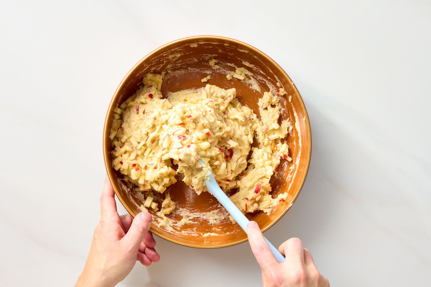 Hands mixing apple fritter batter with a spatula in a bowl
