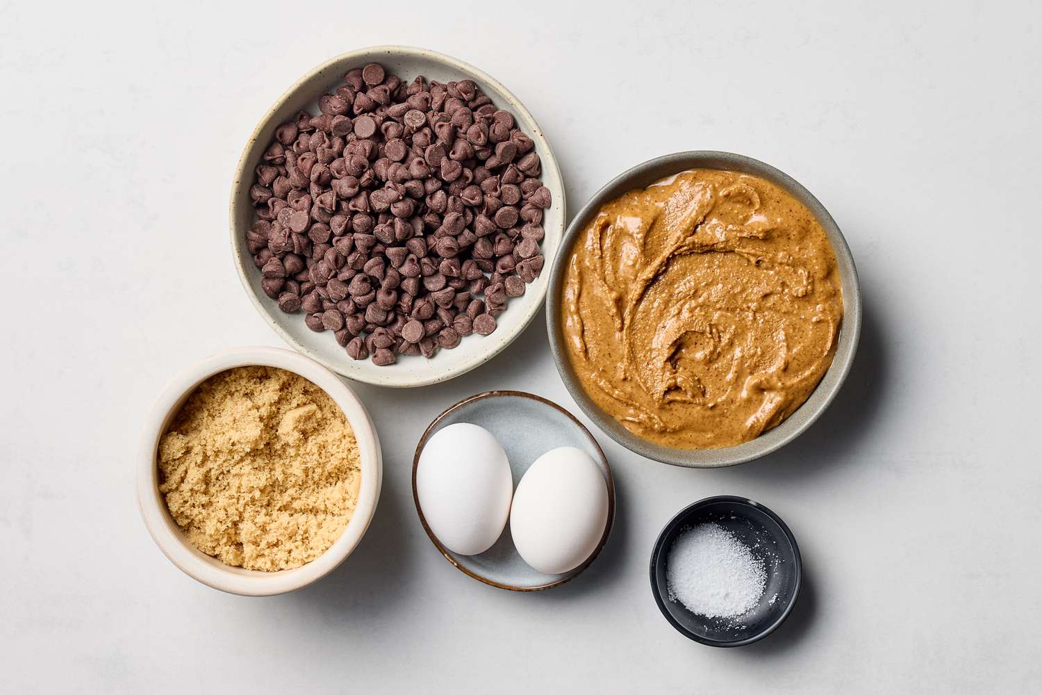 Ingredients for cookies including chocolate chips creamy substance brown sugar two eggs and coarse salt in bowls