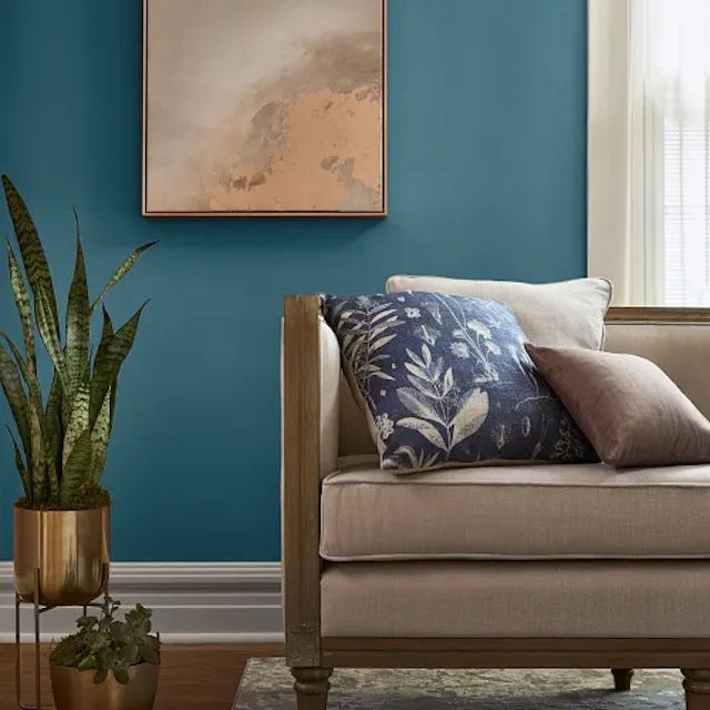A room with a beige couch decorative pillows a blue wall a potted plant and abstract artwork on the wall