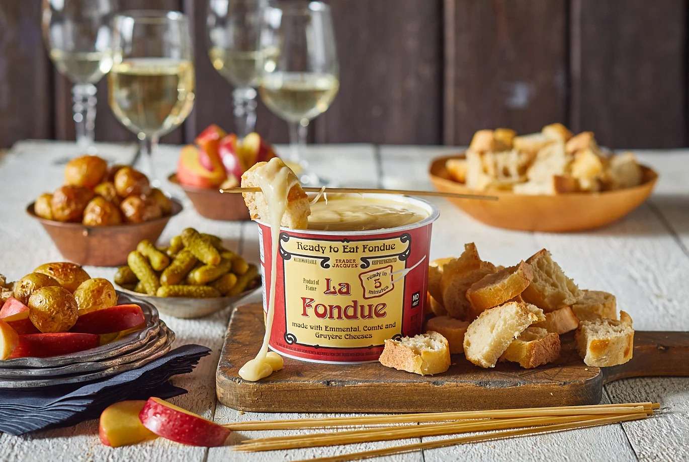 Prepared fondue with bread pickles apples and wine glasses on a table