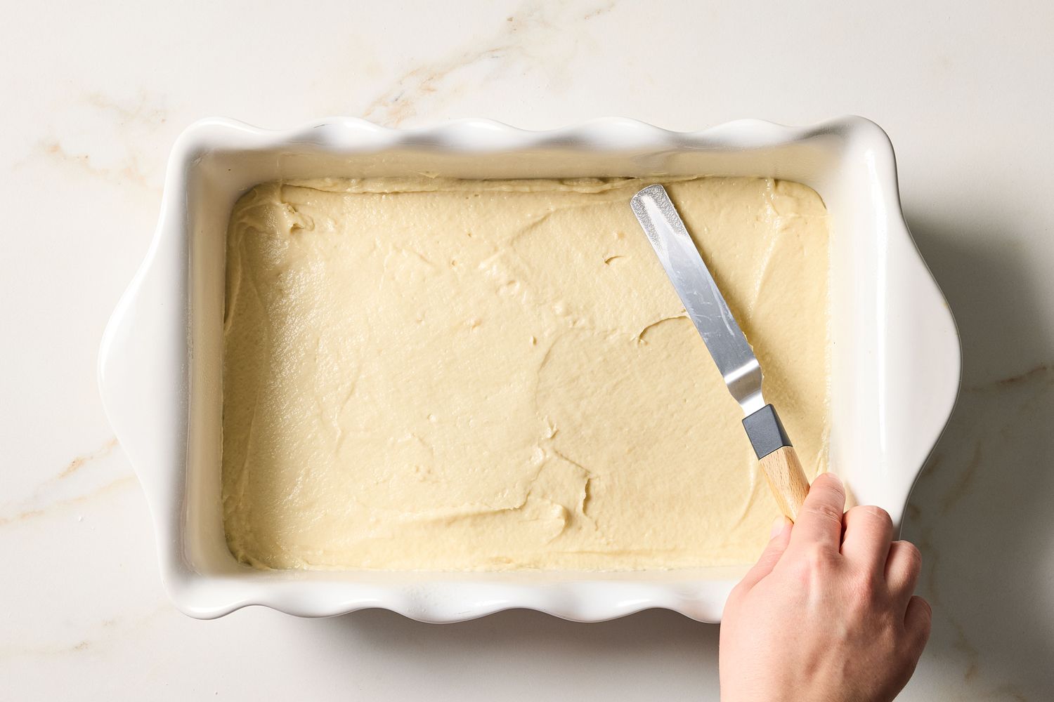 A hand spreading cake batter in a rectangular pan with an offset spatula
