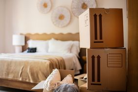 Cardboard boxes stacked in a bedroom with a bed visible in the background
