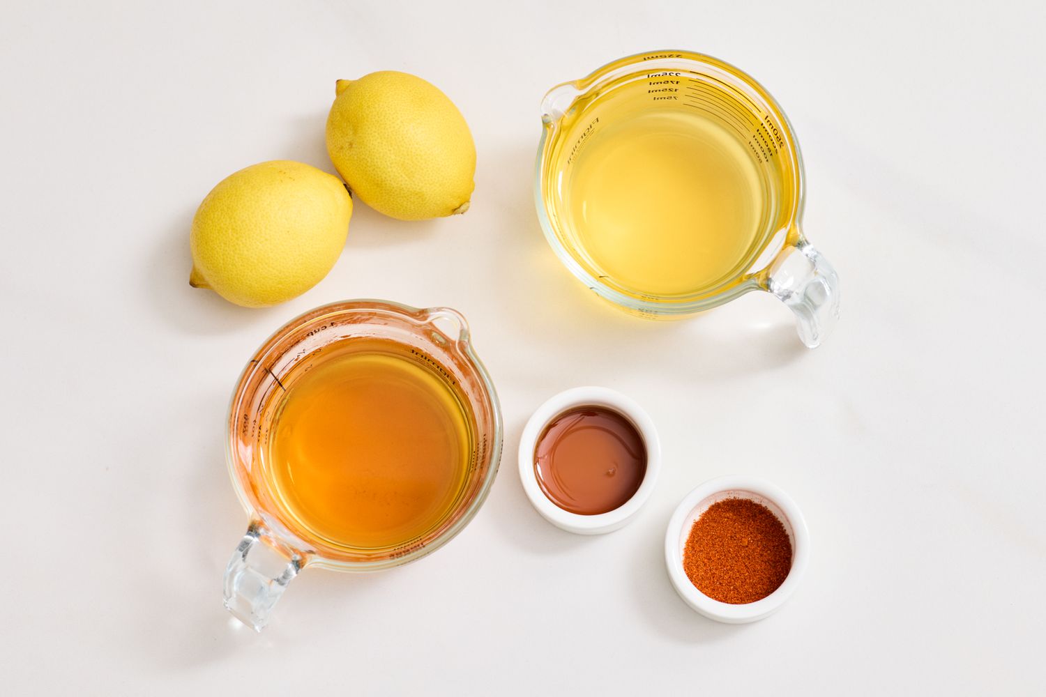 Ingredients including lemons two measuring cups containing liquids and two small bowls with seasonings arranged on a surface