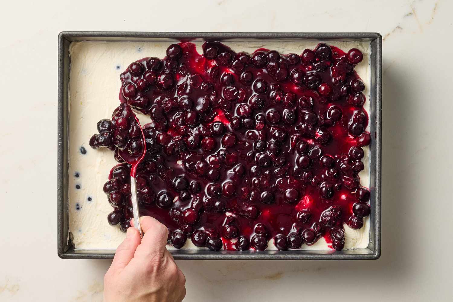 Preparing a ricotta and blueberry cheesecake in a square pan with a hand smoothing blueberry topping using a spoon