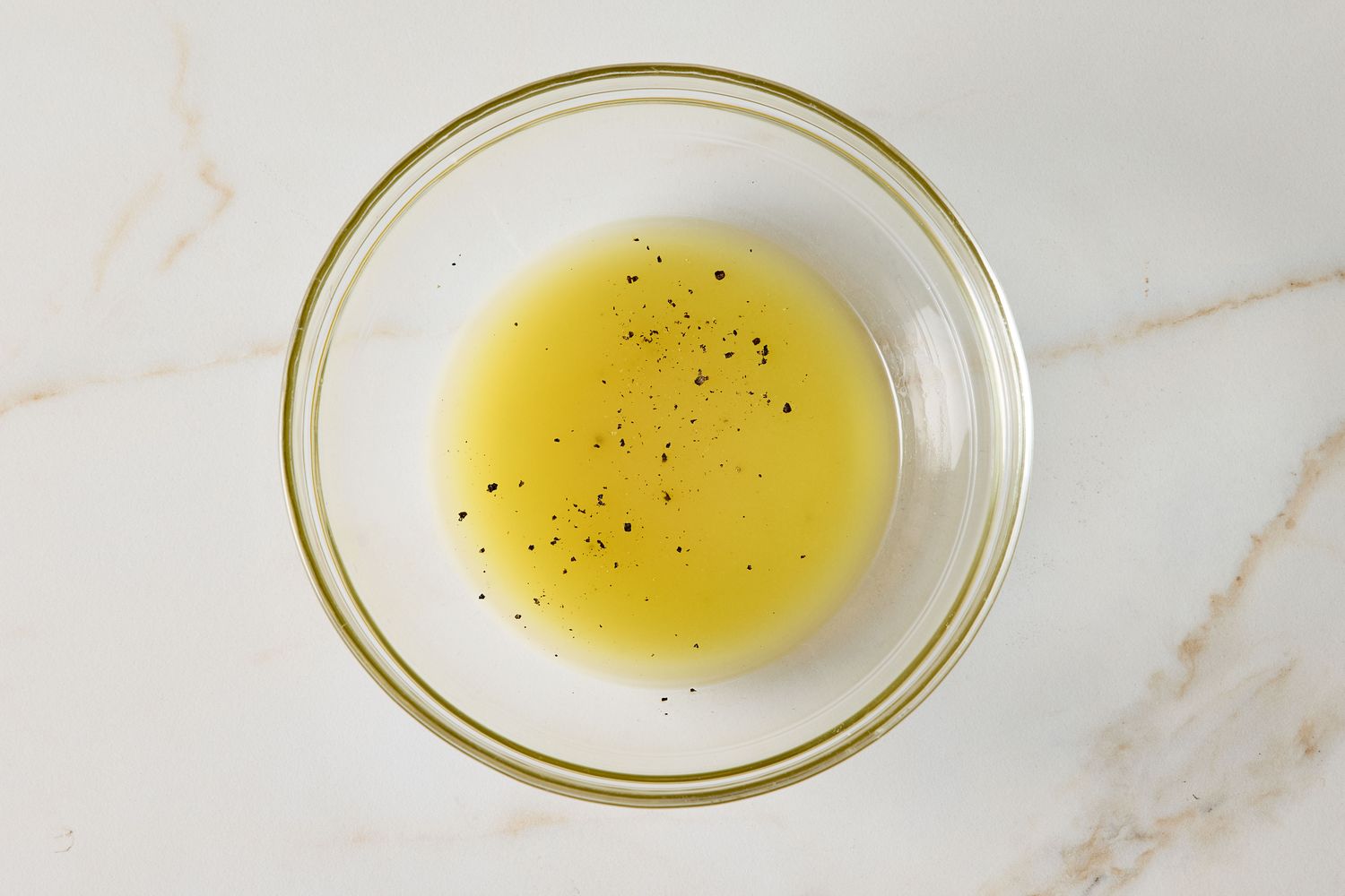A clear glass bowl with liquid and seasoning on a white surface