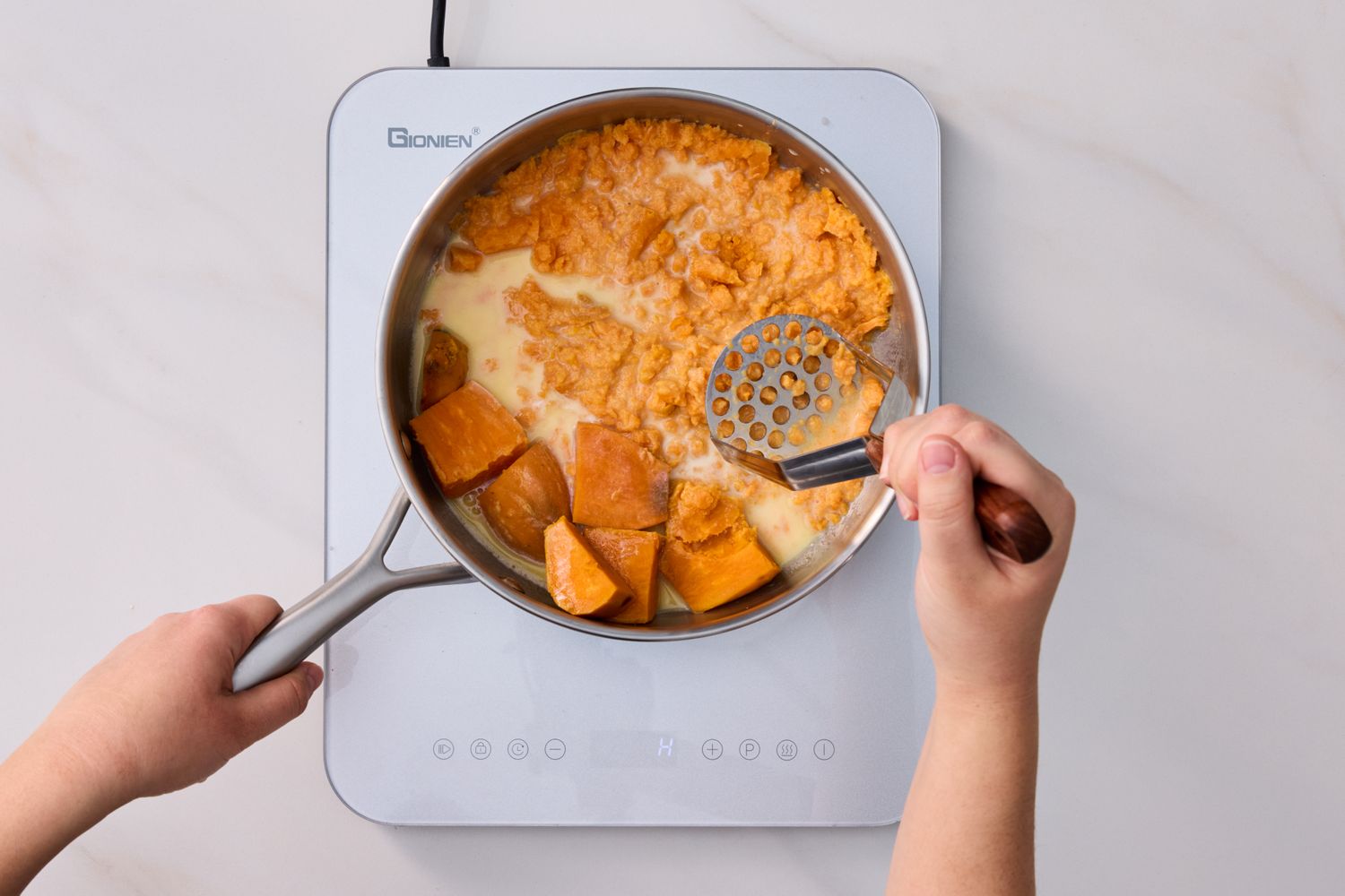 Hands mashing sweet potatoes in a pot on a stove
