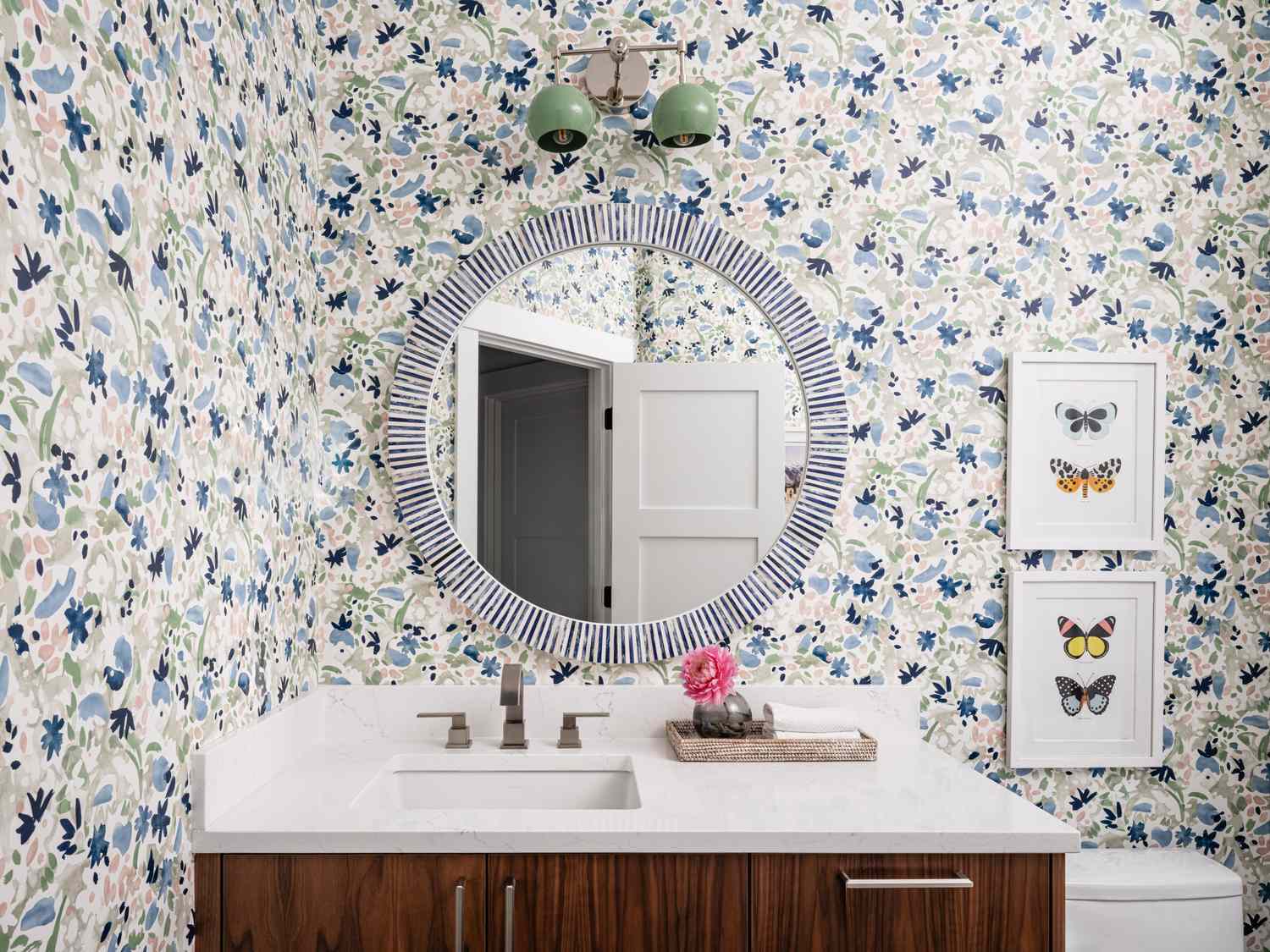 Floral wallpapered bathroom with circular mirror framed butterfly artworks and modern vanity sink area