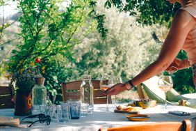 Table set outdoors with various drinks and dishes person pouring wine into a glass surrounded by greenery