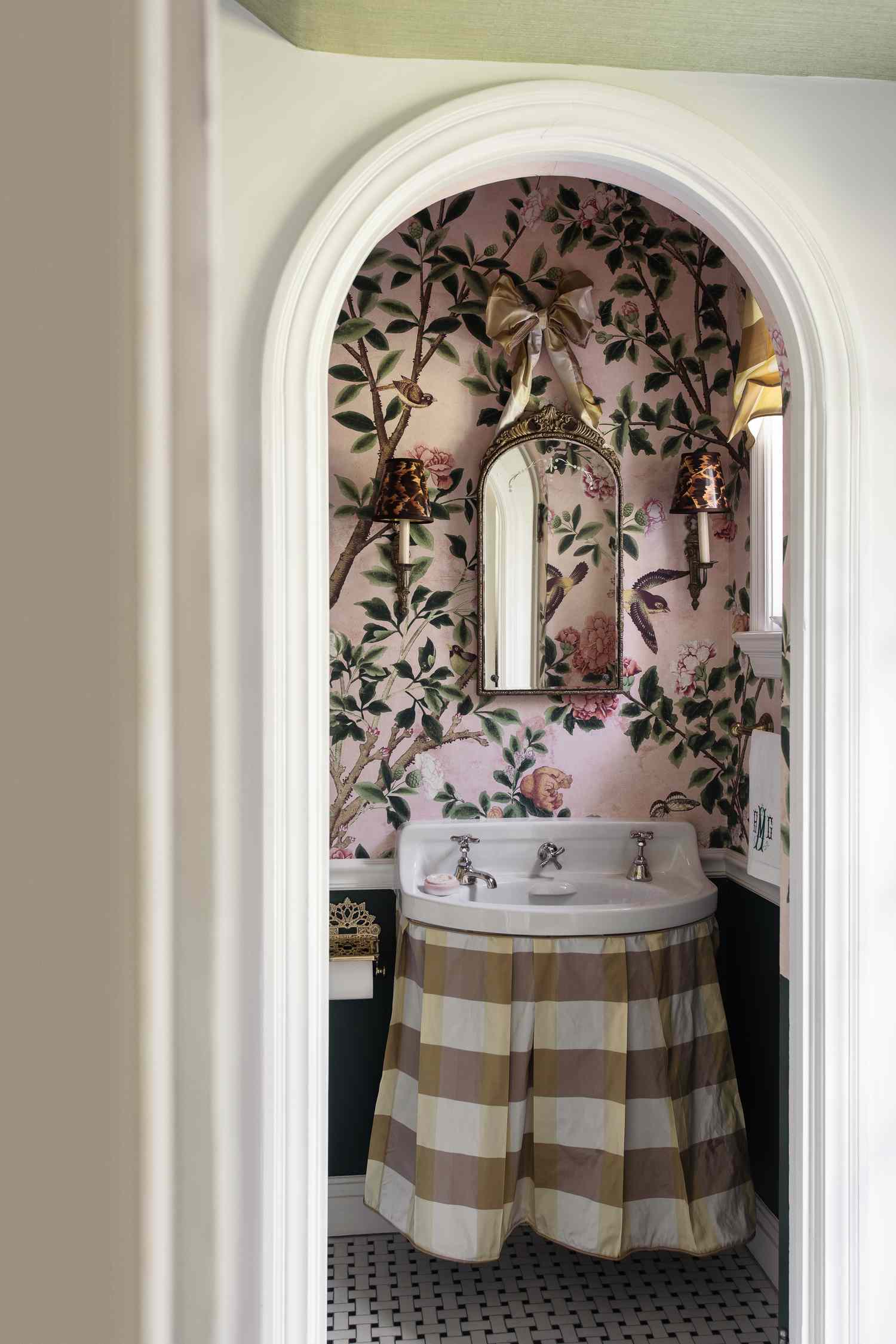 A small bathroom with floral wallpaper an arched doorway a mirror and a fabric-covered sink with a checkered skirt