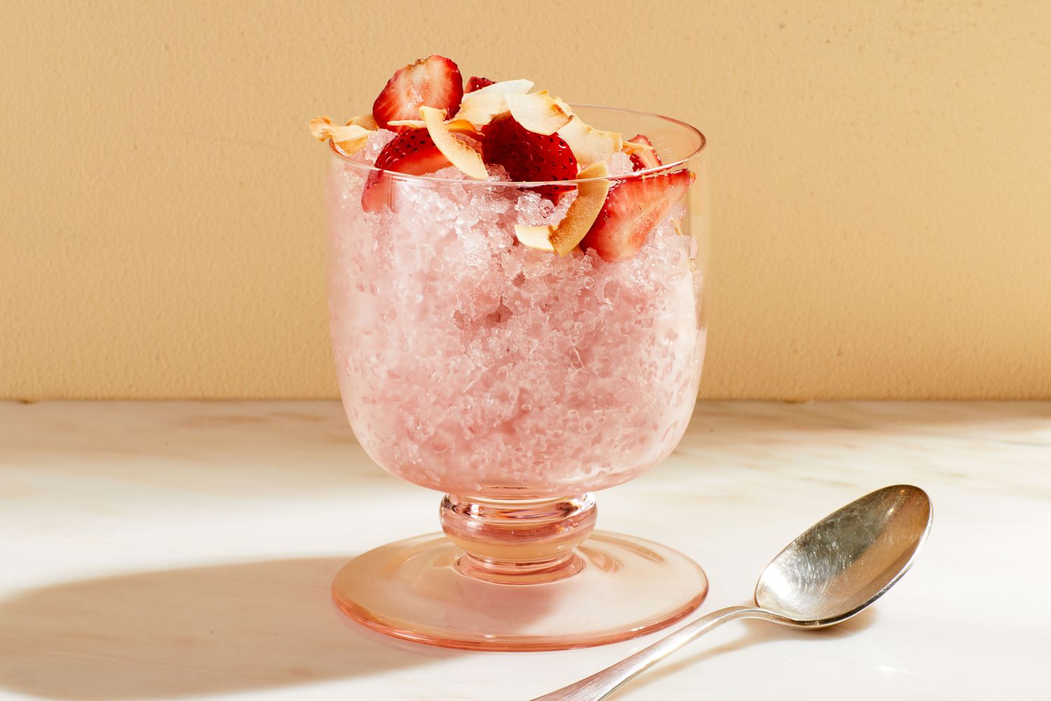 A glass of coconut granita topped with fresh fruit and a spoon beside it
