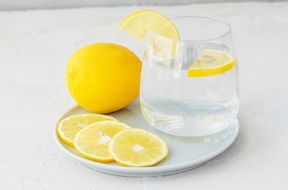 A glass of water with lemon slices on a white plate accompanied by additional lemon slices and a whole lemon