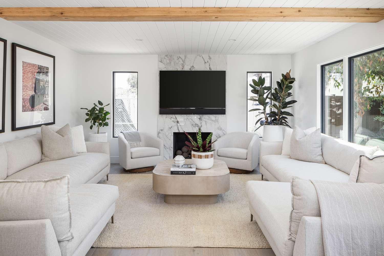 Minimalist living room by Lulu Designs: beige sectional sofas and swivel chairs with pillows, marble fireplace with TV, potted plants, and large windows. Light wood beams, neutral rug, and natural light create a calm, elegant space.
