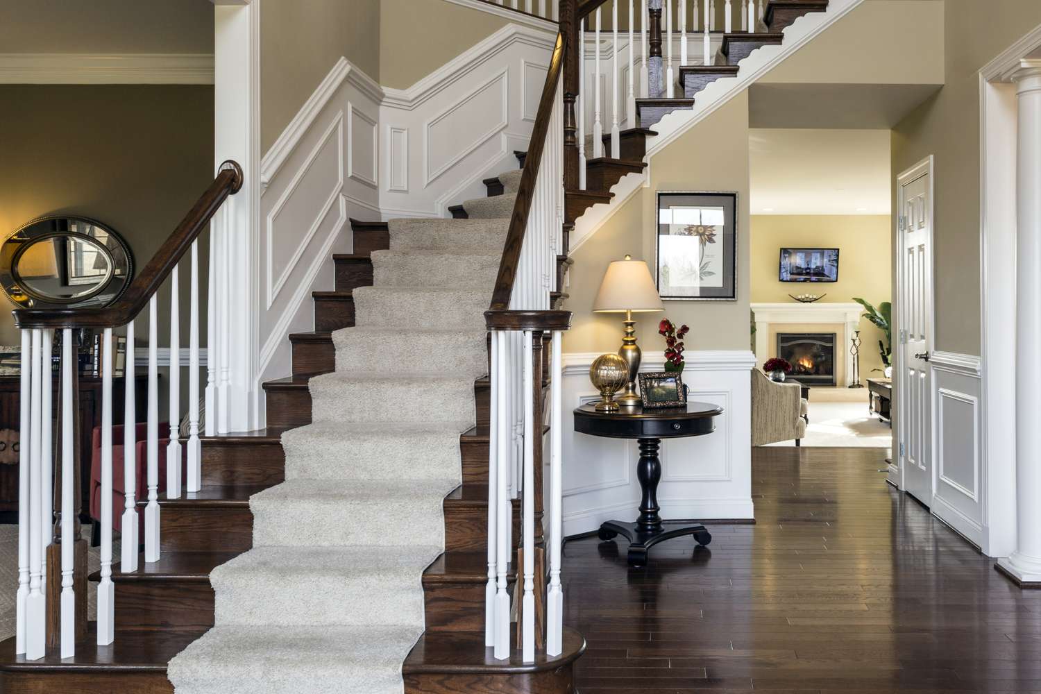 A home interior featuring a staircase with carpeting on steps and wooden handrails a table with a lamp nearby and part of a living room
