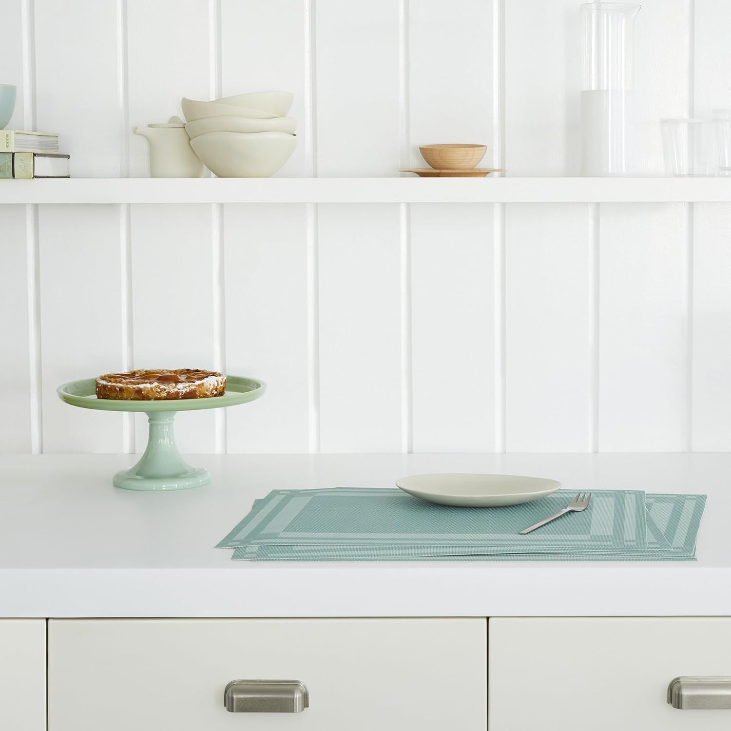 A kitchen counter with a pie on a green cake stand a plate and cutlery on a placemat and open shelves with dishes in the background