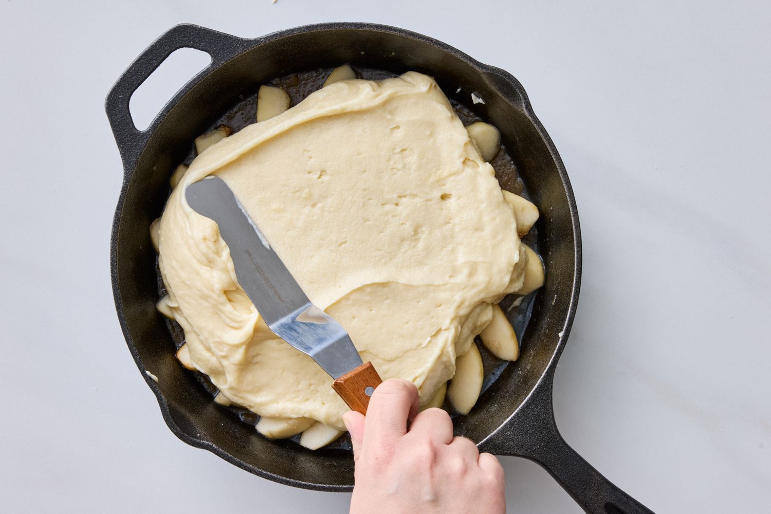 A hand using a spatula to spread cake batter in a castiron skillet containing sliced pears