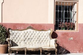 An ornate sofa on a sidewalk against a rustic wall with a small window adorned with plants