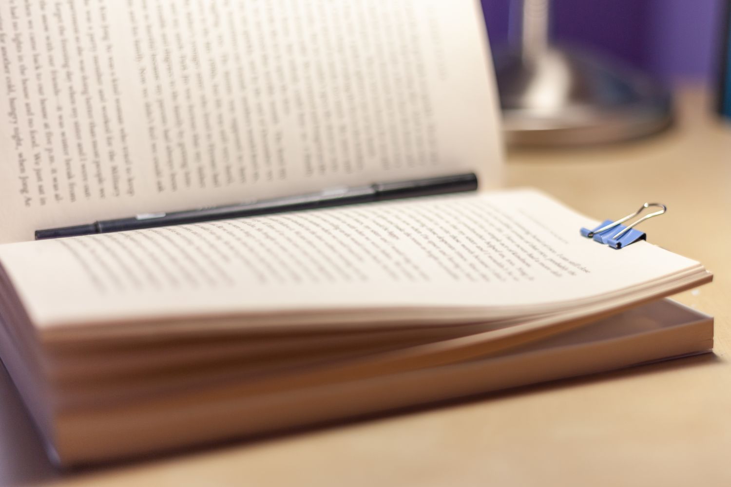 An open book with a binder clip and a pen resting across its pages on a desk near a lamp