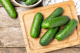A wooden cutting board with several cucumbers placed on it additional cucumbers in a bowl in the background table surface underneath