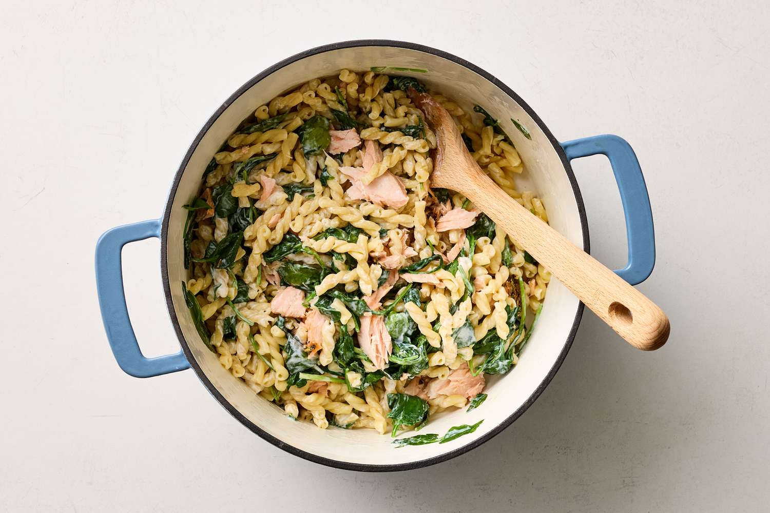 A pot containing cooked pasta mixed with pieces of salmon and greens with a wooden spoon on one side of the pot