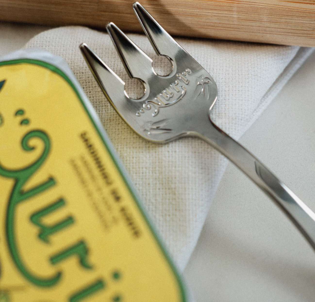 A fish-shaped fork beside a can of Nuri labeled sardines on a white napkin with a wooden element in the background