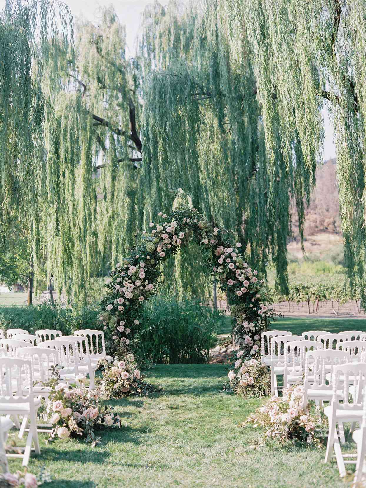 large greenery and pink floral wedding arch in front of willow tree