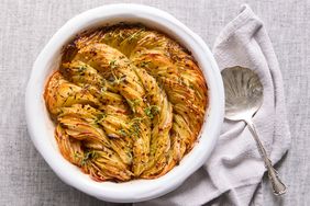 A roasted potato dish baked in a circular pattern in a white dish garnished with herbs served with a metal spoon on the side
