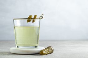 A glass filled with a lightcolored liquid garnished with pickles on a skewer and a pickle on the surface next to it